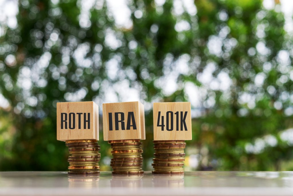 Three stacks of coins with wood blocks on them that have the types of savings accounts, Roth, IRA, and 401k, printed on the wood blocks.