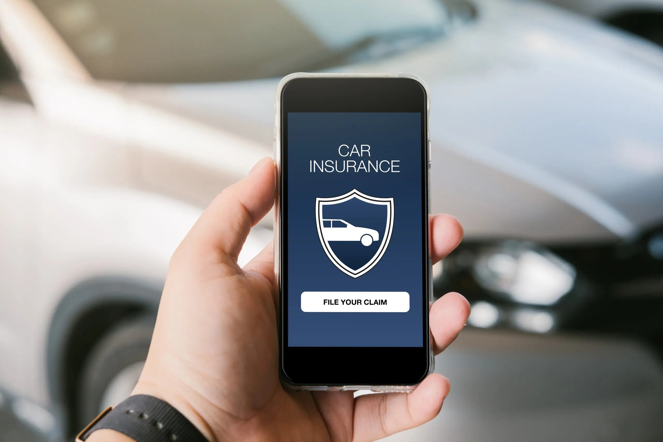 A person holds their phone in frame with a mockup of a car insurance app on the screen. A car is out of focus in the background.