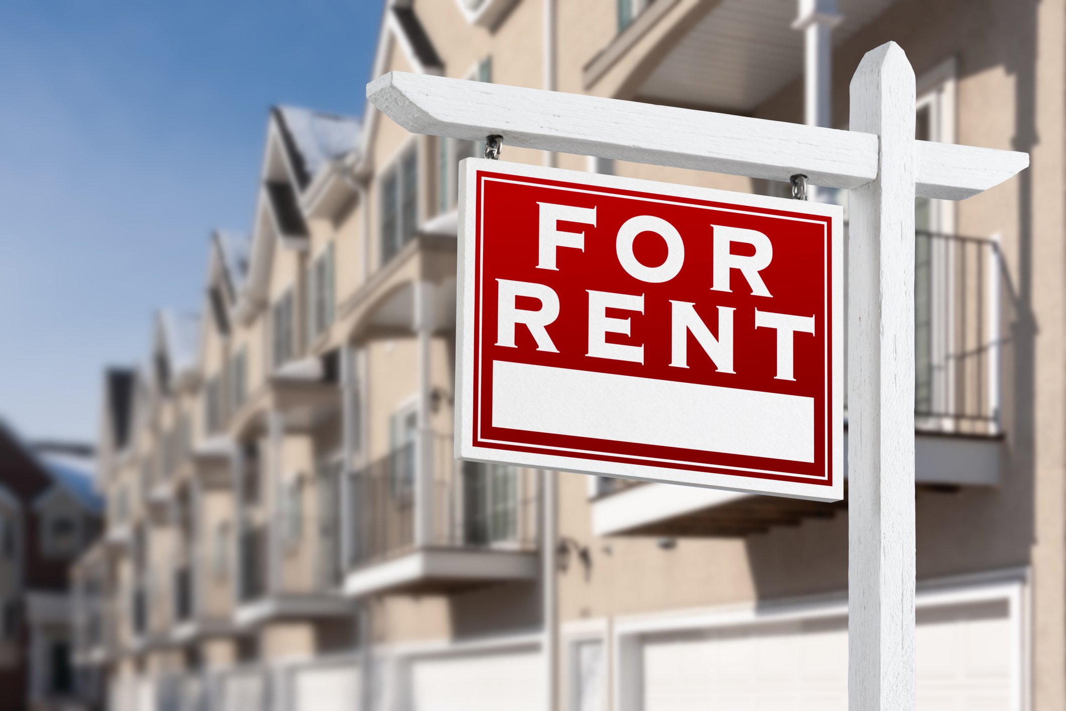 A “for rent” sign stands before a row of beige townhomes.