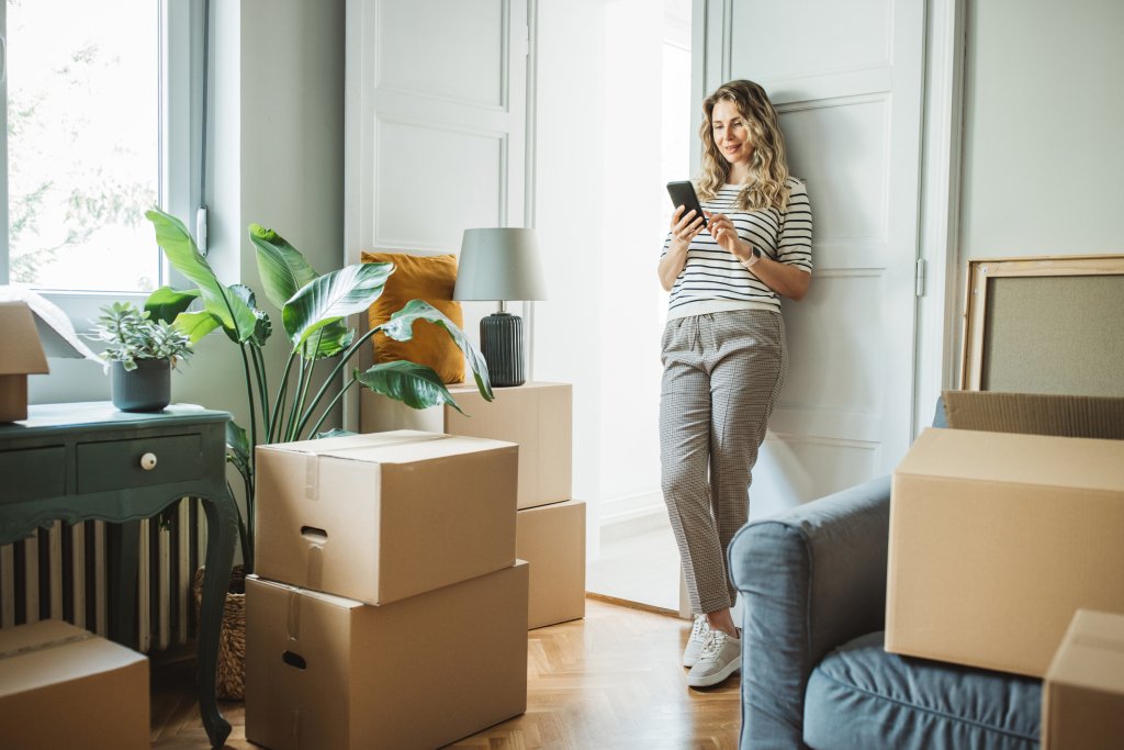 A young woman relaxes in her new apartment. Cardboard boxes are stacked around her, indicating she is in the process of moving in. Lush plants sit by the windowsill.