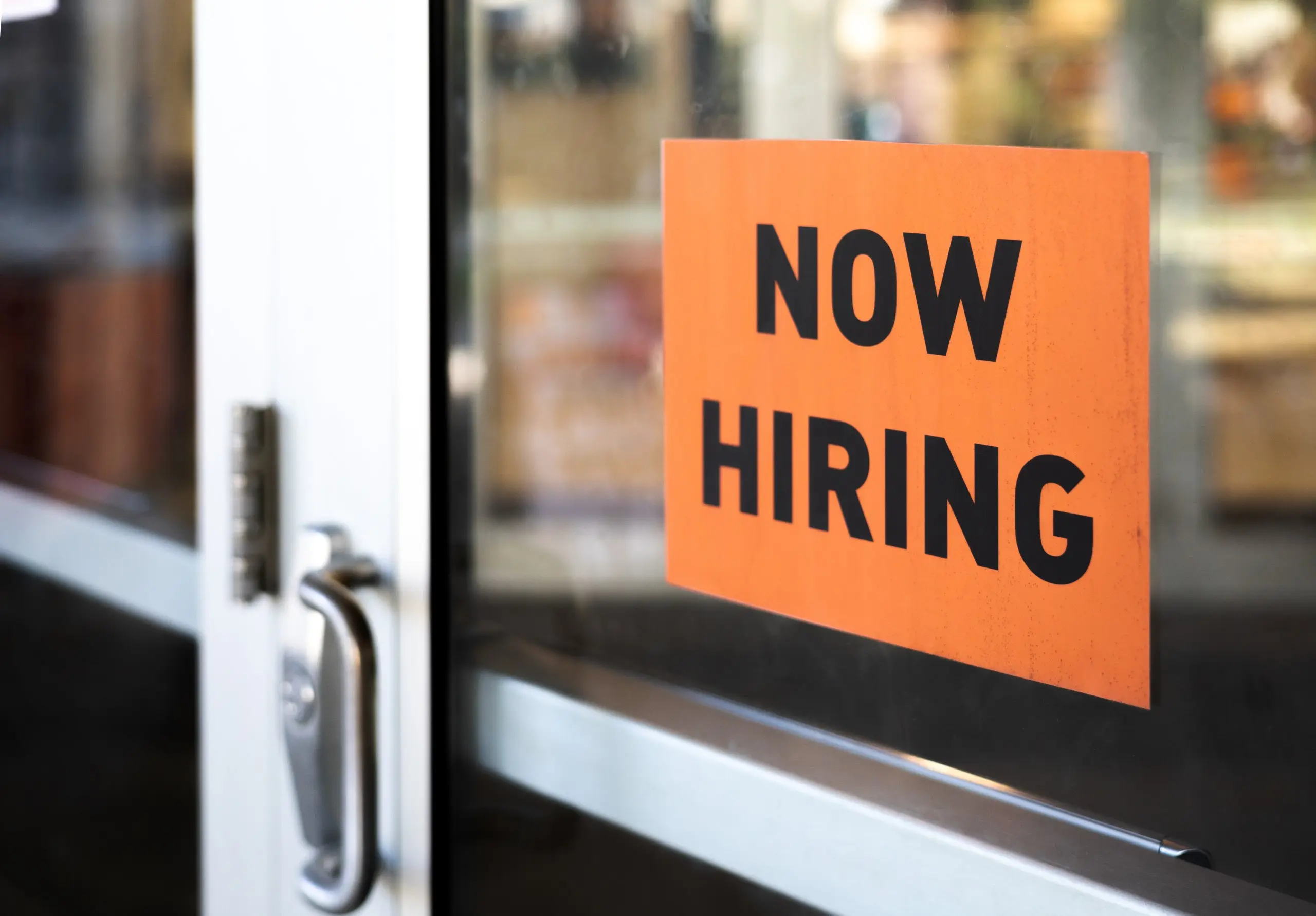 An orange “now hiring” sign is adhered to a glass storefront door.