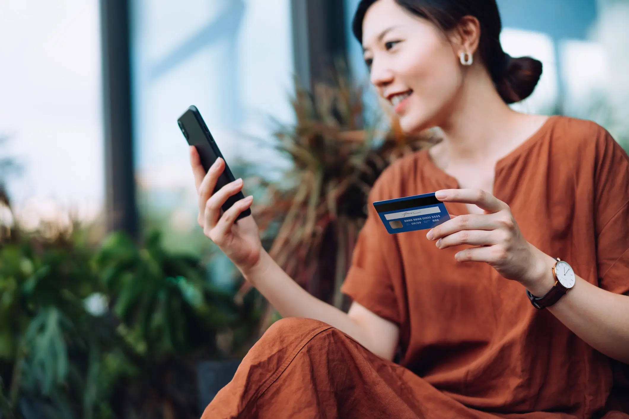 Woman sitting down holding her credit card while typing in her card information on her cell phone.