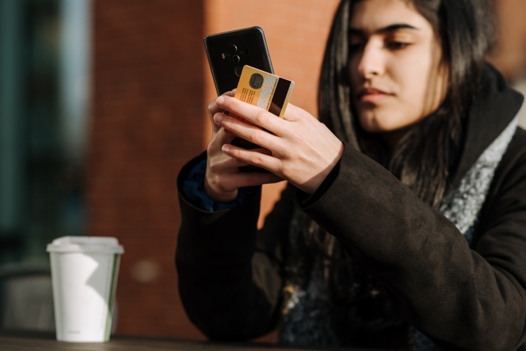 A woman holding her credit card in her left hand uses her cell phone to look for a loan to consolidate her credit card debt.