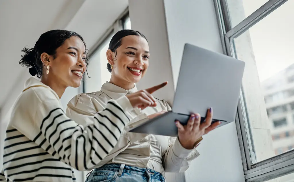 Two young women look at a laptop while smiling. One woman is holding the laptop, and the other is pointing at it as they discuss gig jobs.
