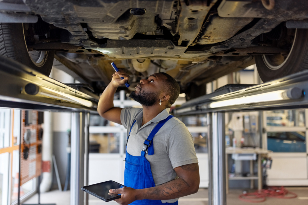 A mechanic looks at the undercarriage of a vehicle with a flashlight while holding a tablet. Used car repair costs can make some purchases more trouble than they’re worth.