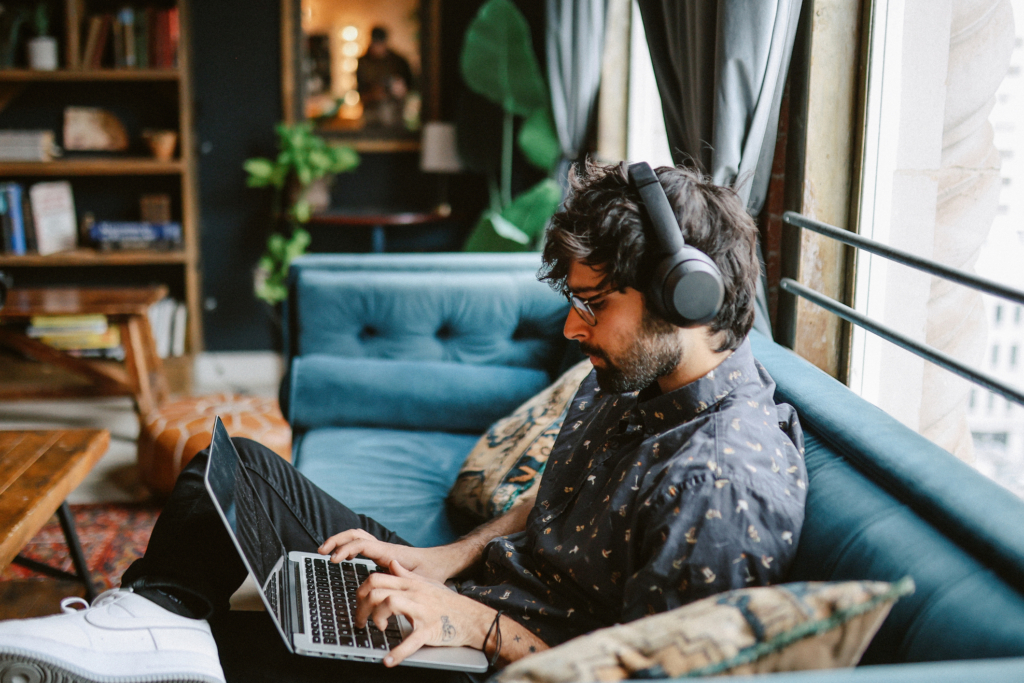 A freelance writer picks up gig work, typing on his laptop in his living room while wearing headphones.