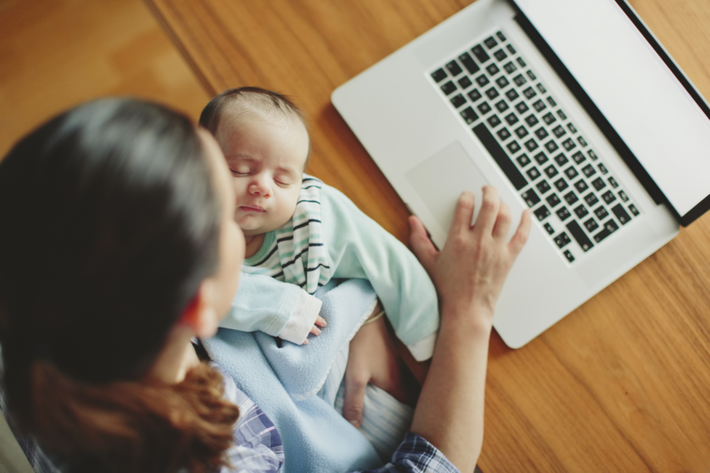 A young woman types on her laptop, looking for gig work to help with childcare expenses while holding a baby.