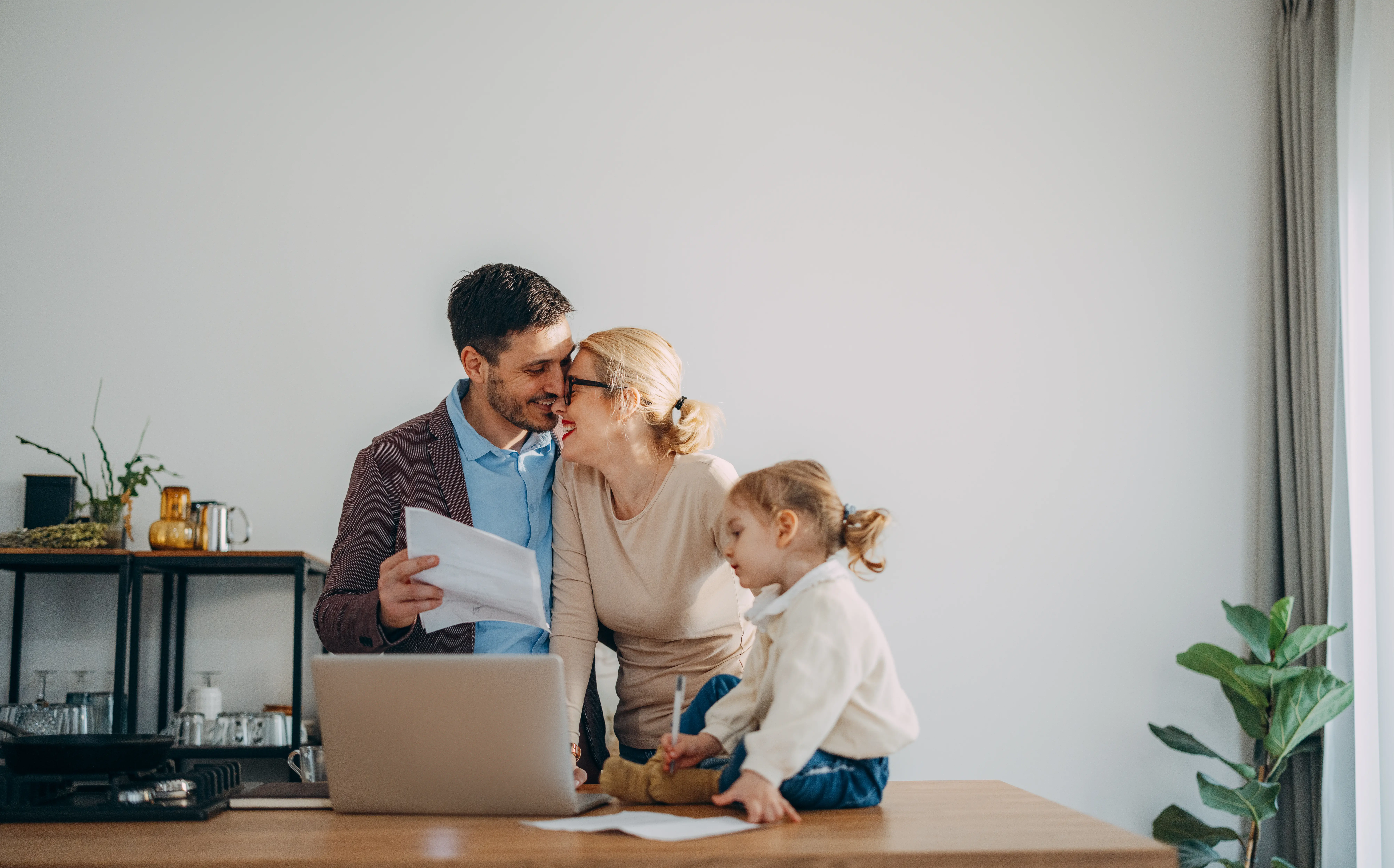 Family standing behind a computer desk with mom and dad hugging, and daughter sitting on the desk