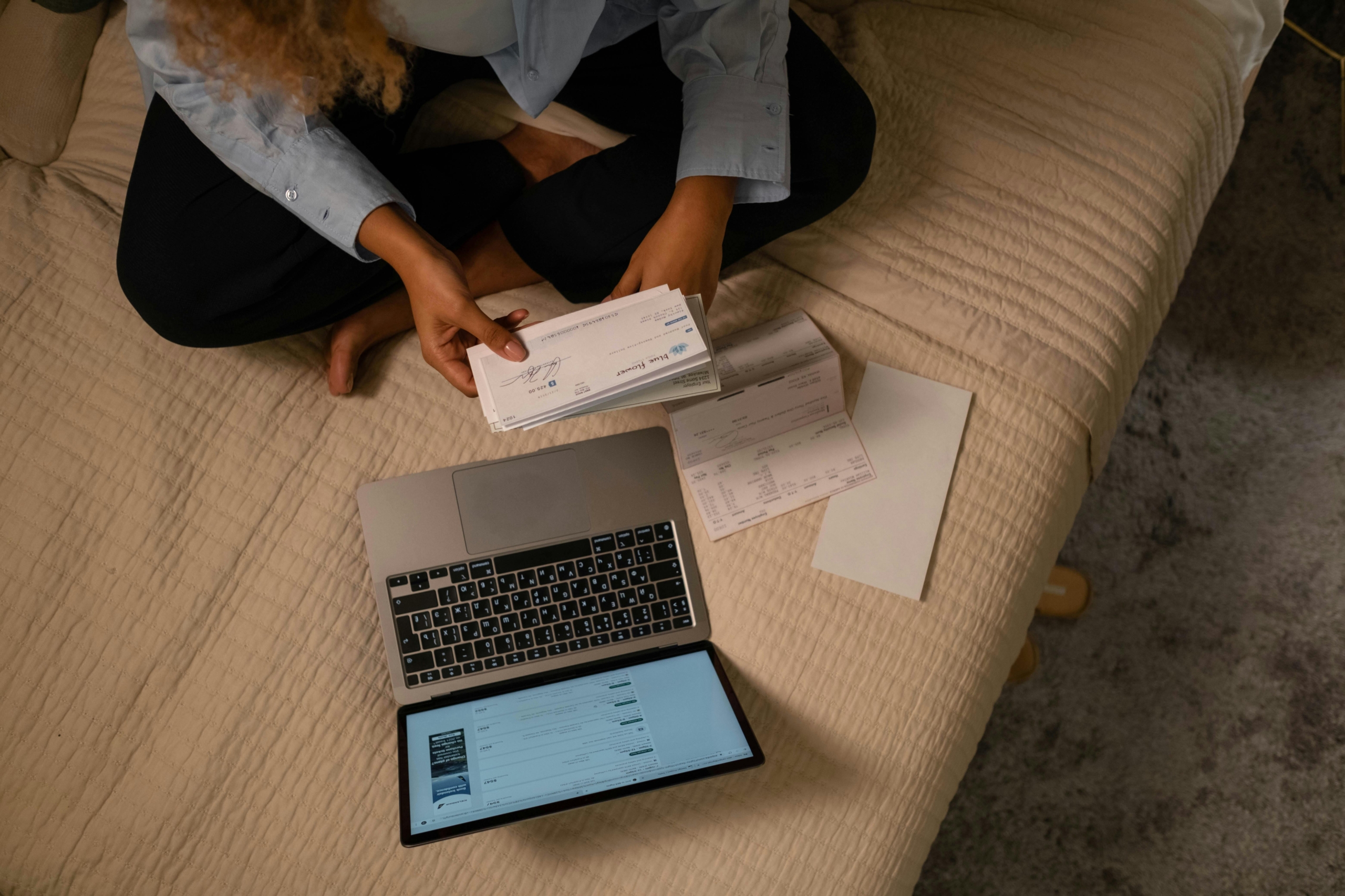 Person sitting on a bed reviewing paychecks and bills next to a laptop