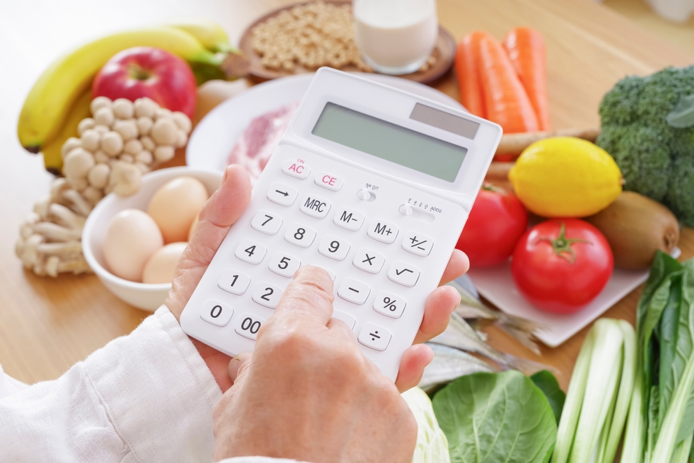 A person using a calculator in front of a table of vegetables.